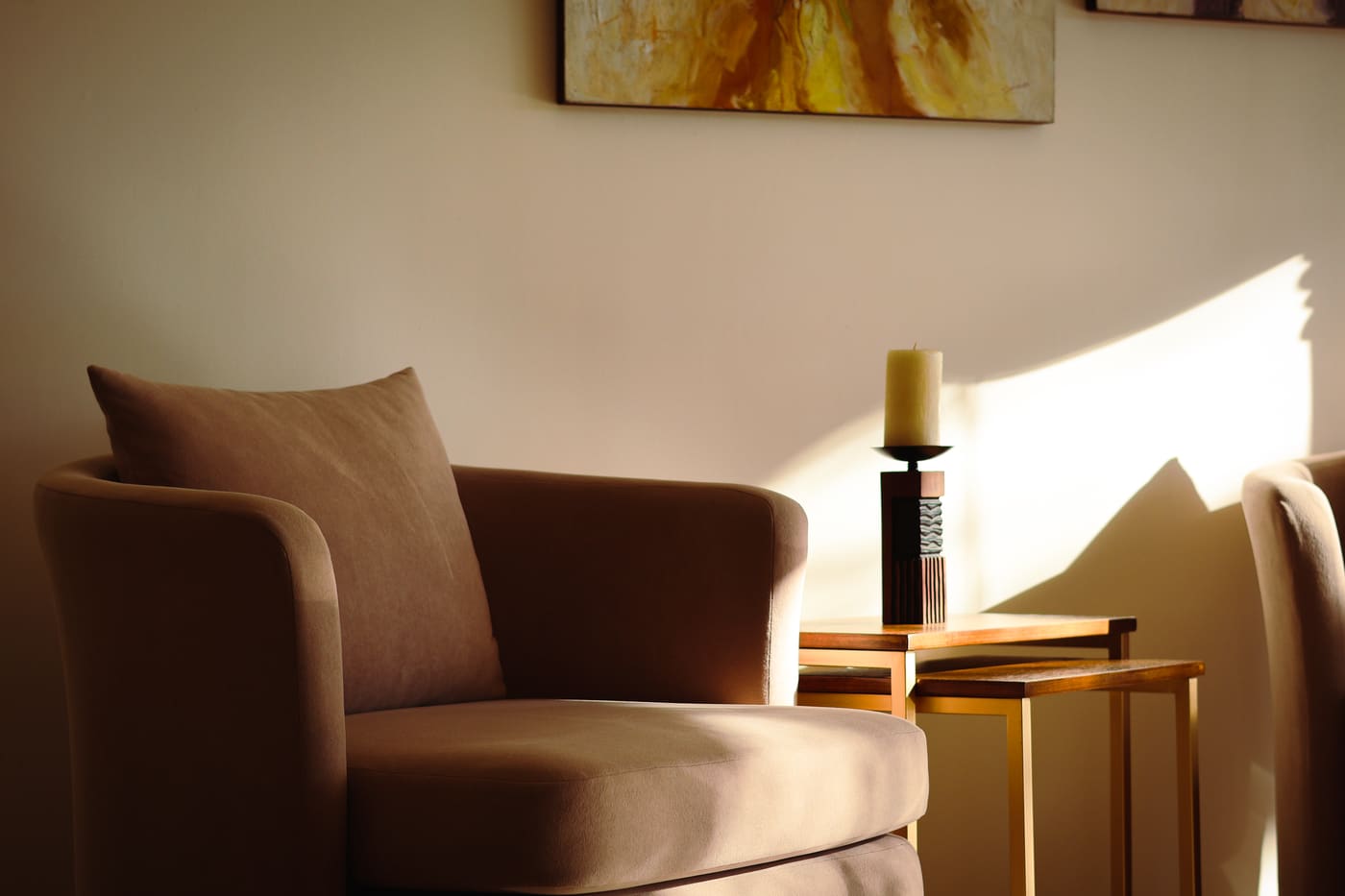 A beige armchair and a lit candle on a wooden table in a sunlit corner